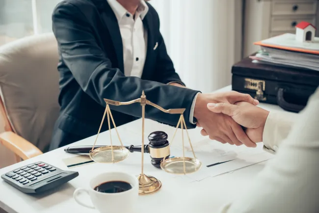 Lawyer shaking hands with client in office with legal balance scale and gavel on desk.